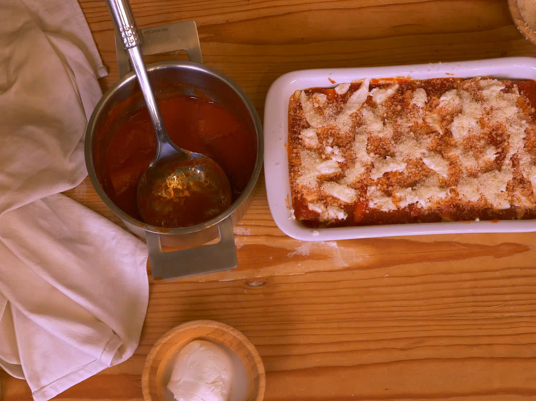 A baking dish of meat cannelloni under tomato passata and Parmigiano, next to a pot of sauce and fresh mozzarella.