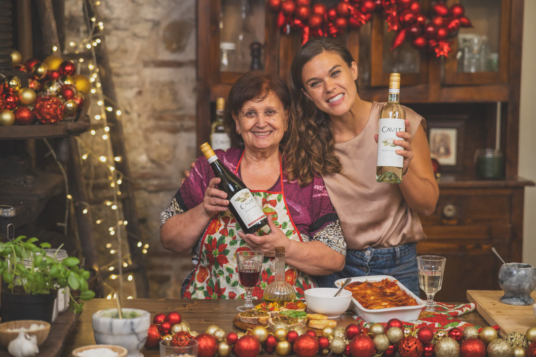 Nonna holds a bottle of Cavit Pinot Noir and the host holds Cavit Pinot Grigio behind the finished cannelloni and bruschetta.