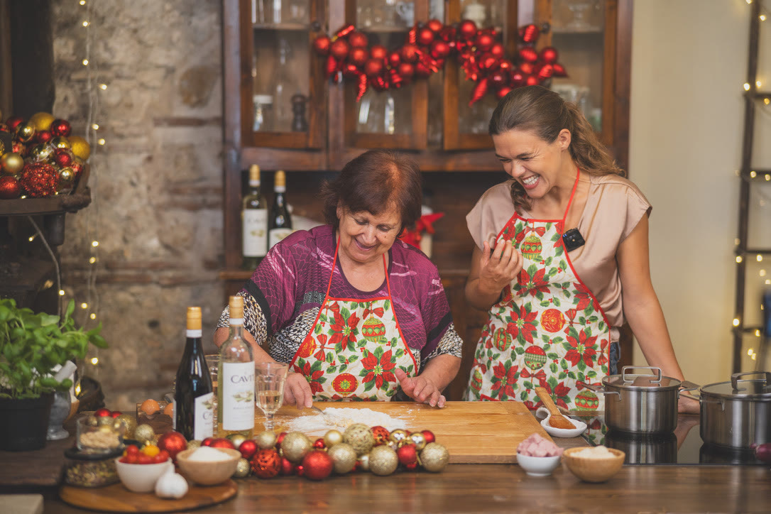 Nonna smiles as she works flour into fresh pasta dough while the class host laughs beside her, with bottles of Cavit on the counter.