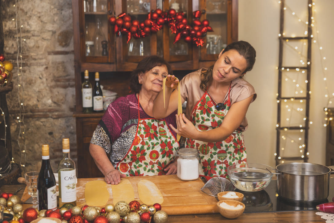 The host holds up a thin, translucent strip of fresh pasta as Nonna looks on approvingly.